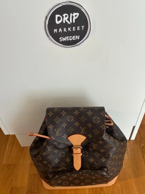 Brun monogram ryggsäck - Snygg brun ryggsäck i klassiskt brunt monogrammönster med beigea och bruna läderdetaljer. Ryggsäcken har ett lock med spänne, dragkedjeficka framtill och justerbara axelband. Perfekt för dig som vill ha en lyxig och trendig accessoar.