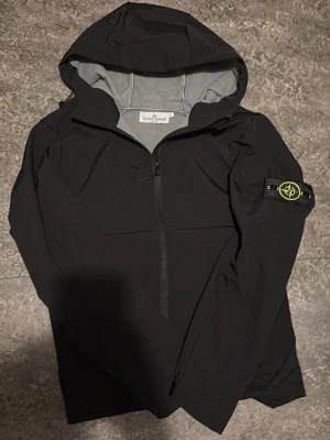 Svart windbreaker med huva Stone Island - Svart vindjacka från Stone Island med huva och ikonisk patch på vänster ärm. Jackan har half zip framtill och är gjord i ett lätt, syntetiskt material. Perfekt för dig som gillar stilrena och funktionella plagg.
