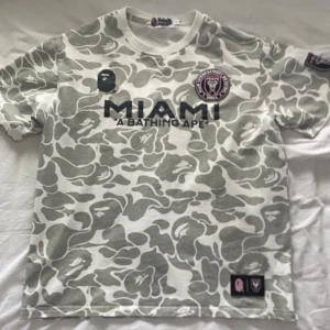BAPE T shirt - Säljer en unik vit och grå camo t-shirt från MIAMI x A Bathing Ape med tryck både fram och bak. Stora BAPE-loggan och siffran 93 på ryggen. Klassisk rund hals och korta ärmar. Perfekt för dig som gillar streetwear och vill sticka ut. Passar Mer M än L