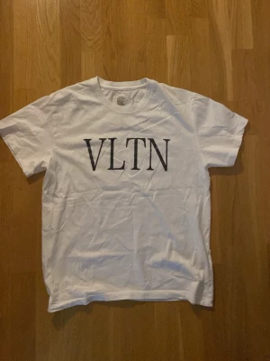 Vit Valentino t-shirt med tryck - Säljer en vit t-shirt med svart VLTN-tryck på bröstet. Klassisk rund hals och korta ärmar. Perfekt basic-plagg med stilren design som funkar till det mesta. Tillverkad i mjuk bomull för skön känsla.