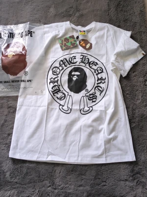 Vit BAPE x Chrome Hearts T-shirt - Säljer en vit T-shirt från samarbetet mellan BAPE och Chrome Hearts. Framsidan har ett stort svart tryck med BAPE-loggan och Chrome Hearts-typografi, medan baksidan har Baby Milo och ett korsmotiv. Klassisk passform och rund hals. Perfekt för dig som gillar streetwear.