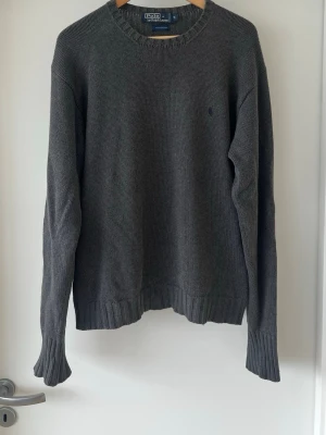 Mörkgrå stickad tröja Polo Ralph Lauren - Klassisk mörkgrå stickad tröja från Polo Ralph Lauren med rund halsringning och diskret broderad logga på bröstet. Tröjan har långa ärmar och ribbade muddar vid ärmslut och nederkant. Perfekt basplagg i garderoben. Lappen i nacken har lossnat på ena sidan, annars bra skick.