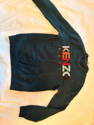 Mörkgrön stickad Kenzo-tröja - Snygg mörkgrön stickad tröja från Kenzo med färgglad logga broderad framtill. Tröjan har rund halsringning och ribbade muddar vid ärmslut och nederkant. Perfekt för dig som vill ha en trendig och bekväm look.