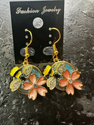 Blommiga örhängen med blommor och bin - Unika örhängen med stora rosa blommor, glittrande stenar och ett glittrande bi som detaljer. Basen är turkosblå. Hängande modell med krok i guldfärg. Perfekt för dig som gillar färg och statement-accessoarer.