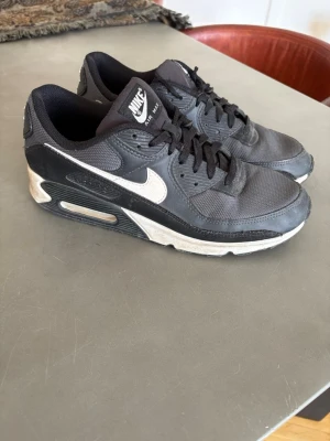 Nike Air Max svarta sneakers - Säljer ett par klassiska Nike Air Max sneakers i svart med vit sula och vit swoosh. Skorna har en låg profil, mesh och syntetmaterial på ovandelen samt synlig Air Max-dämpning i sulan. Perfekta för dig som gillar streetwear och sportig stil.