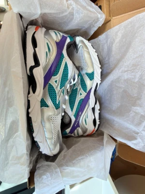 Mizuno Wave Rider 10 sneakers - Snygga Mizuno Wave Rider 10 sneakers i vitt, turkos och lila med meshpaneler och detaljer i syntet. Skorna har en chunky sula och cool retrostil med Mizuno-logga på sidan. Perfekta för dig som gillar sportig och trendig look.