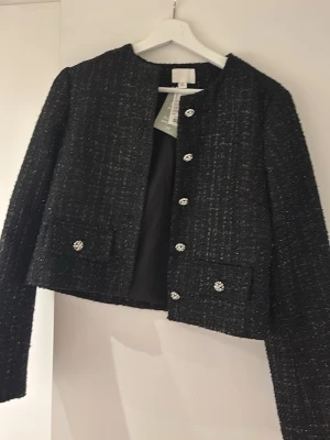 Svart tweedkavaj med glittriga knappar - Stilren svart tweedkavaj från H&M med glittriga, dekorativa knappar framtill och på fickorna. Kavajen har rak passform, rund halsringning och är fodrad. Materialet är återvunnen polyester och ger en exklusiv känsla. Perfekt för att lyfta din outfit.