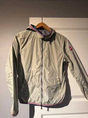 Beige vindjacka med huva - Snygg beige och grå colmar vindjacka med huva och dragkedja framtill. Jackan har röda och blå detaljer på krage och nederkant samt en rund logotyp på ärmen. Tillverkad i lätt och tunt material, perfekt för blåsiga dagar. Två fickor med dragkedja på sidorna. Jackan har nypris på 2700kr