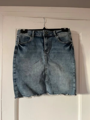 Blå jeanskjol med rå kant - Snygg blå jeanskjol i klassisk modell med fem fickor och bälteshällor. Kjolen har en rå, fransig kant nedtill för en trendig look och stängs med knapp och dragkedja framtill. Perfekt att matcha med en enkel t-shirt eller hoodie.