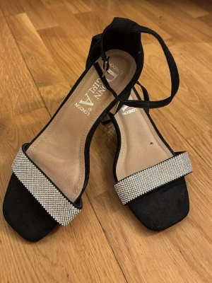Svarta pumps med strass från Donna Girl - Snygga svarta pumps från Donna Girl med fyrkantig tå och bred rem framtill dekorerad med glittrande strass. Skorna har en smal ankelrem med spänne och en elegant, öppen design. Perfekta för att lyfta vilken outfit som helst.