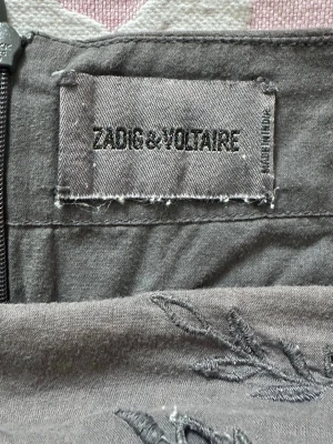 Grå broderad kjol från Zadig & Voltaire - Snygg grå kjol från Zadig & Voltaire i 100% bomull med broderade detaljer. Kjolen har dragkedja i sidan och ett stilrent uttryck med diskreta broderier som ger en unik touch. Perfekt för dig som gillar minimalistisk men ändå personlig stil.