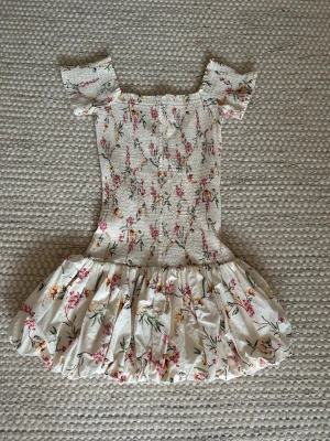 Ny slutsåld Kort blommig klänning med volang från H&M  - Söt kort klänning i ljus botten med blommigt mönster i rosa, gul och grön. Klänningen har puffiga korta ärmar, smockad överdel och volangig kjol. Perfekt för sommarens alla tillfällen och ger en romantisk vibe.