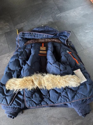 Parajumper righthand parka - Snygg mörkblå dunjacka med avtagbar päls på huvan och orangea detaljer. Jackan har quiltat mönster, dragkedja och tryckknappar samt en mysig insida. Perfekt för kalla vinterdagar. Skriv om du har några frågor mvh