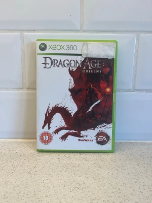 Dragon Age: Origins till Xbox 360 - Dragon Age: Origins till Xbox 360 är ett episkt fantasyrollspel från BioWare och EA. Omslaget är vitt med röd drake och mörka illustrationer. Spelet är i fysisk form med plastfodral och PEGI 18-märkning. Perfekt för dig som gillar djupa berättelser och strategi . Fungerar super