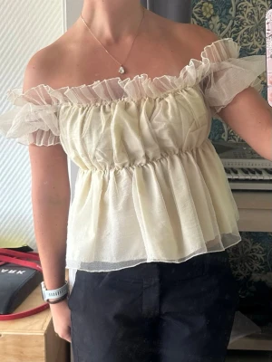 Offshoulder volangtopp ljusgul - Söt offshoulder topp i ljusgult med volangdetaljer och transparenta lager. Toppen har en luftig passform och resår vid axlar och midja för en feminin siluett. Perfekt på festen eller utgången med tjejerna!