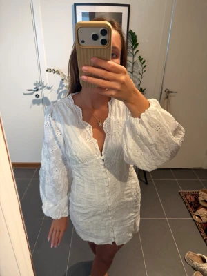 Vit kort klänning med spetsdetaljer - Söt vit kort klänning med broderade spetsdetaljer och knappar framtill. Klänningen har långärmade puffärmar och v-ringad halsringning med dekorativ kant. Perfekt för sommarens alla tillfällen.