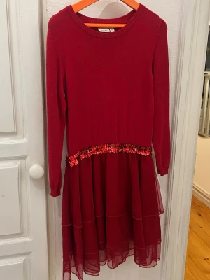 Röd klänning med paljettdetalj - Röd långärmad klänning med rund halsringning och volangkjol i flera lager. Midjan är dekorerad med röda paljetter som ger en extra touch. Kjolen har ett skirt, lätt tyg som ger ett fint fall. Från märket name it, storlek 128 (inga defekter) 💖
