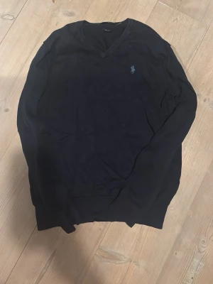 Mörkblå V-ringad tröja Polo Ralph Lauren - Klassisk mörkblå V-ringad tröja från Polo Ralph Lauren med den ikoniska broderade loggan på bröstet. Tröjan har långa ärmar och ribbade muddar vid ärmslut och nederkant. Perfekt för en stilren och avslappnad look.