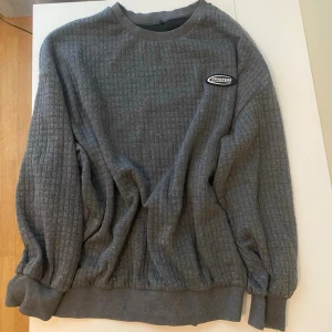 Grå ribbad långärmad topp - Säljer en grå långärmad topp med ribbad struktur och rund hals. Toppen har en diskret patch på bröstet och muddar vid ärmslut och nederkant. Perfekt för en chill och avslappnad stil.