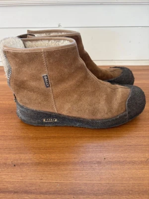 Bruna mockastövlar från Bally - Bruna tofflor i mocka med mjukt, ljust foder och kraftig svart sula. Tofflorna har en enkel, rak design och dragkedja baktill. Perfekta för dig som vill ha både värme och stil hemma. En klassiker som håller dig varm om fötterna. Sulan har börjat släppa pyttelite i tån. Bara att fixa hos skomakaren.  Skorna är använda men har många år kvar.  Strl 37.