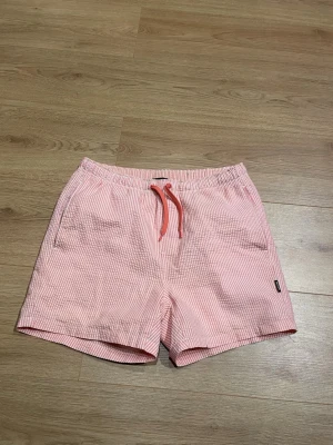 Randiga rosa badshorts Only & Sons - Säljer ett par snygga badshorts från Only & Sons i ljusrosa och vitt med tunna ränder. Shortsen har elastisk midja med snörning och sidofickor. Perfekta för sommaren och strandhäng.