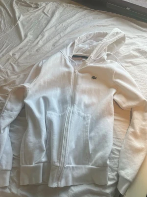 Vit zip hoodie från Lacoste - Snygg vit hoodie från Lacoste med dragkedja framtill och klassisk krokodil-logga på bröstet. Hoodien har huva med dragsko, två fickor och ribbade muddar vid ärmar och nederkant. Perfekt för en clean och sportig look.