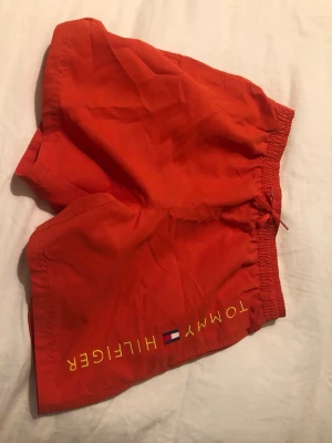 Oranga badshorts Tommy Hilfiger - Snygga oranga badshorts från Tommy Hilfiger med gul logotyp och flagga på ena benet. Shortsen har elastisk midja med dragsko och en bakficka med knapp. Perfekta för stranden eller poolen.