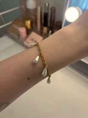 Guldigt armband med droppformade pärlor - Snyggt armband i guldfärgad metall med hängande droppformade pärlor. Kedjan är tunn och smidig, och pärlorna ger en elegant touch. Perfekt accessoar för att lyfta din outfit. Handgjort