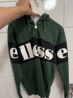 Grön Ellesse hoodie med dragkedja - Snygg grön hoodie från Ellesse med stor vit logga över bröstet och svarta paneler. Tröjan har huva med dragsko och halv dragkedja framtill. Tillverkad i mjukt bomullsmaterial, perfekt för en avslappnad streetwear-look.