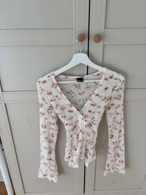 Vit blommig omlottopp med knyt - Supersöt vit omlottopp med små blommor i orange och grönt. Toppen har lång ärm, v-ringning och knyt i ryggen. Ribbad struktur som ger en skön och stretchig känsla. Perfekt för dig som gillar romantisk och trendig stil.