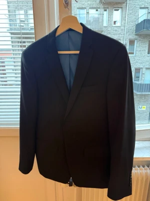 Svart kavaj från Erla of Sweden - Stilren svart kavaj från Erla of Sweden med klassisk passform och blått innerfoder. Kavajen har två innerfickor med orange detaljer och en bröstficka. Perfekt för dig som vill ha en tidlös och snygg look.