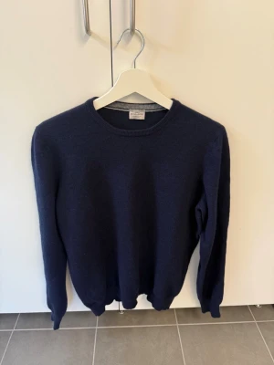 Mörkblå stickad tröja Rose & Born, stl M/50 - Klassisk mörkblå stickad tröja från Rose & Born. Tröjan har rund halsringning, långa ärmar och ribbade muddar vid ärmslut och nederkant. Perfekt för lager på lager och enkel att matcha med olika stilar. Tillverkad i Ull, Kashmir och Viskose.