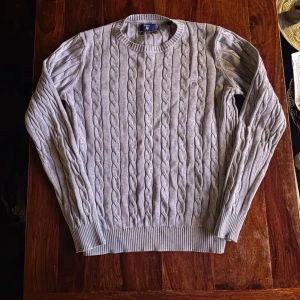 Ljusgrå Gant kabelstickad tröja - Snygg ljusgrå kabelstickad tröja ifrån GANT med rund halsringning och ribbade muddar vid ärmslut och nederkant. Tröjan har ett diskret broderat emblem på bröstet och är perfekt för lager på lager. Klassisk och tidlös design i mjukt material.