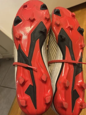 Grå och röda fotbollsskor med dobbar - Säljer ett par grå fotbollsskor med röda detaljer och svarta ränder på sidan. Skorna har röda snören, stickad strumpliknande öppning och röda dobbar undertill för bra grepp på planen. Perfekta för dig som vill ha stilrena och funktionella skor till matchen.