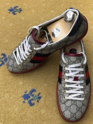 Gucci sneakers GG supreme Canvas  - Snygga Gucci sneakers i beige canvas med klassiskt GG-mönster, röda och marinblå ränder på sidan samt röda detaljer vid hälen. Skorna har vita skosnören, guldfärgade öljetter och röd sula. Perfekta för dig som vill sticka ut med exklusiv stil.