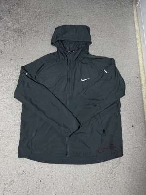 Svart Nike vindjacka med huva - Snygg svart vindjacka från Nike med huva och hel dragkedja framtill. Jackan har långa ärmar med resår vid handlederna och reflekterande detaljer på ärmarna samt Nike-logga på bröstet. Tillverkad i lätt och vindtätt material, perfekt för blåsiga dagar.