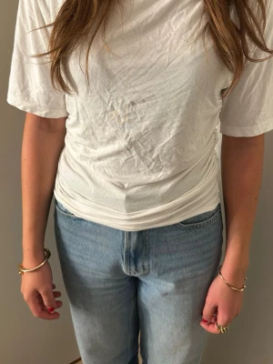Vit t-shirt med draperad midja - Säljer en vit t-shirt med korta ärmar och draperad effekt i midjan. T-shirten har en något lös passform upptill och smalnar av vid midjan, vilket ger en snygg siluett. Materialet ser ut att vara mjuk bomull eller bomullsblandning.
