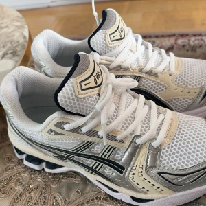 Vita Asics sneakers med mesh och detaljer - Snygga vita sneakers från Asics med mesh-överdel, svarta och silverfärgade detaljer samt beige inslag. Skorna har en sportig design med tydlig logga på sidan och bekväm sula med svart och beige mönster. Perfekta för en trendig och aktiv stil.