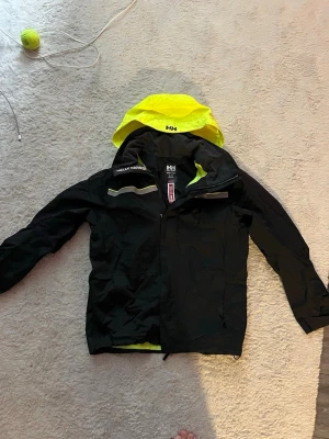 Svart vindjacka Helly Hansen M - Snygg svart vindjacka från Helly Hansen med neongul huva och detaljer. Jackan har dragkedja, reflexdetaljer och regular fit. Perfekt för blåsiga dagar och outdoor-aktiviteter. Materialet är slitstarkt och vattenavvisande.