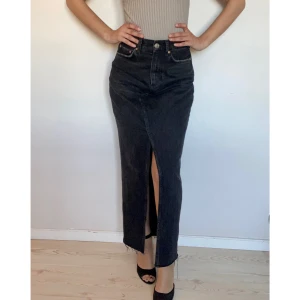 Svart denimkjol  - Svart Denimkjol med knapp och dragkedja i midjan. Slits fram och råa kanter nertill. Omsydd av ett par jeans. Mått:  Längd: 100cm  Midja: 72cm  Höft: 100cm