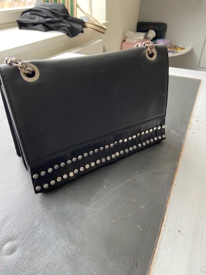 Handväska svart med nitar - Säljer min handväska som jag använt fåtal gånger. Kan ha den på axeln och även justera så den blir längre och ha den korsat över axeln. Den är röd inuti och svart utanpå med fransar och silver kedja.