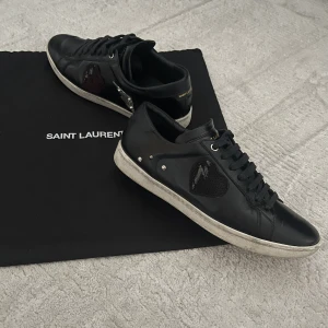 Yves Saint Laurent Sneakers - Säljer dessa svarta glansiga skor från Yves Saint Laurent med detaljer på sidorna (höger fot: rött hjärta och blixt, vänster fot: svart hjärta och nitar). Nypris ligger runt 6000kr.  Storlek 39🙌 Boxen är borta men dustbag medkommer🖤🖤