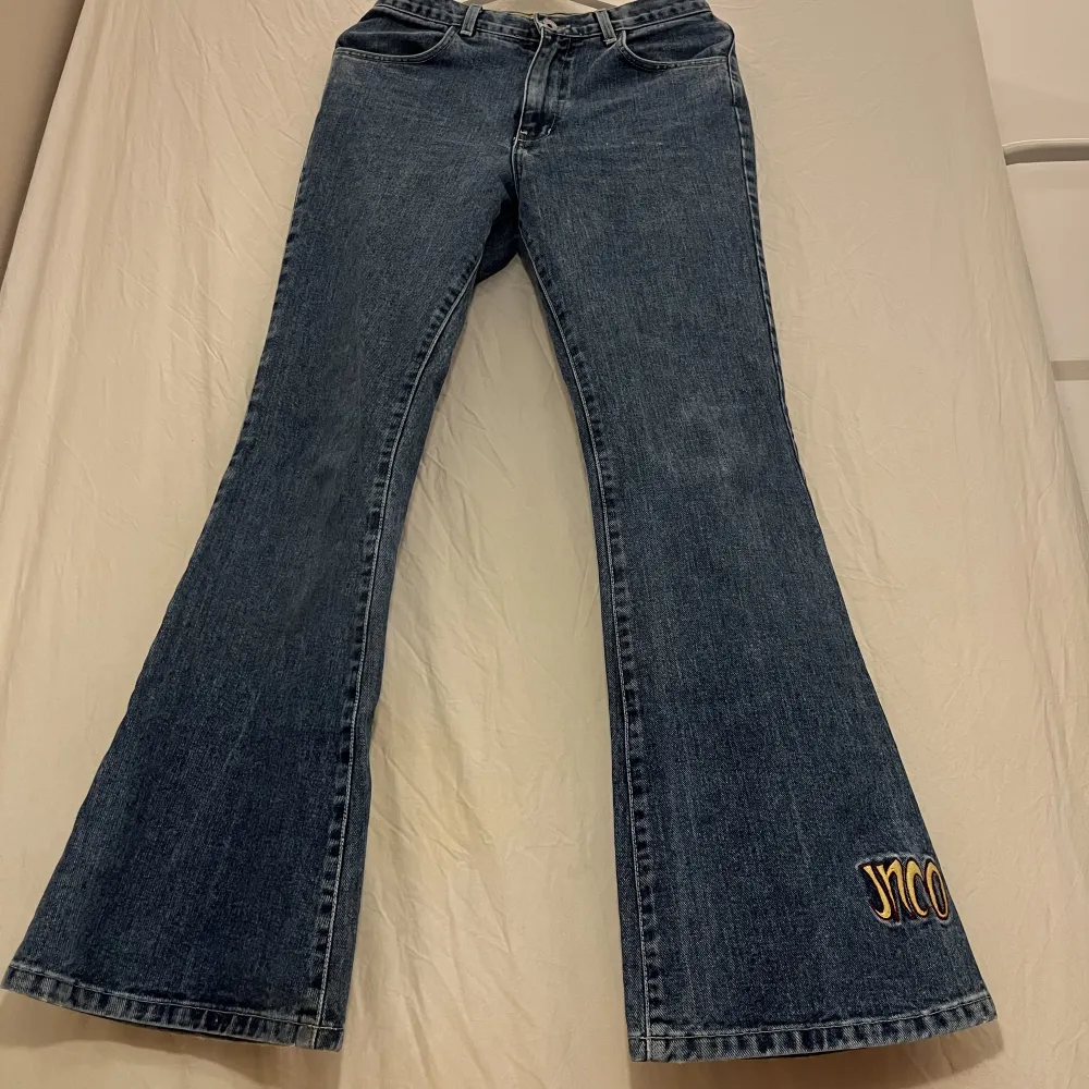 Blå jeans med gul detalj nertill på vänster ben från JNCO. Fint skick. Midmidjade och utsvängda. obs: ungefärliga mått: Midjemått: 78cm. Innerbenslängd: 82cm. . Jeans & Byxor.