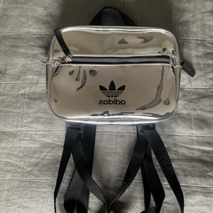 Adidas väska - Silvrig, reflekterande väska från adidas med stort och ett mindre fack. Justerbara band vilket gör att den funkar som både handväska och ryggsäck. Rymlig och lättanvänd. Aldrig använd utan precis som ny🤍