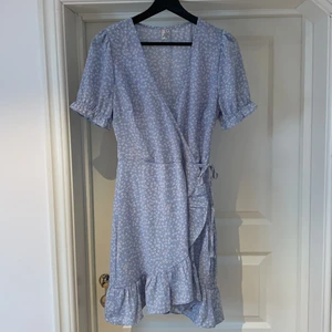 Somrig kort klänning från Nelly, storlek 38 - Jättefin ljusblå omlottklänning från Nelly trend, storlek 38! Söta puffärmar och volang. Röd/vita blommor i mönstret. Använd enstaka gång, nyskick!