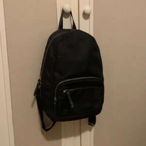 Medelstor svart ryggsäck - Säljer denna gulliga lilla ryggsäck då den inte bara hänger i garderoben! Ej använd och i bra skick! 3 fack och ett innefack i det största facket! Nypris: 299kr