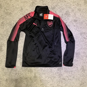 Arsenal träningströja  - En snygg och helt oanvänd Arsenal träningströja från 2017-2018 säsongen med tags kvar. Helt oanvänd och därmed i 10/10 skick. Storlek S men passar nog M också. Den har två fickor med dragkedja och en half zip. Den är äkta. Pris kan diskuteras.