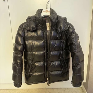 Moncler Maya Down Jacket - Color: Black Condition: 9/10 Size: 0  Features  - Detachable hood  - Two-way front zipper closure  - Elastic drawstring in the hood and bottom hem  - Branded zippers  - Two front pockets  - Moncler badge on left sleeve  - Badge on left sleeve pocket 