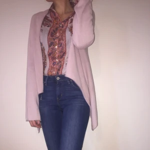  - "Kvadratisk formad cardigan. Essential Japan Knit kan bäras på många olika sätt. Använd den dekorativa nålen för knäppning eller krokarna för att haka fast på önskad plats i tyget. Du kan även bära Essential Japan Knit öppen för en avslappnad look. Detta är en äkta Hunkydory klassiker som funnits i kollektionen sedan varumärket grundades. Bär den över ett enkelt linne eller T-shirt. 100% Bomull"  Storlek S, rosa. Bra skick. Har köpt på Tradera, men används ej.