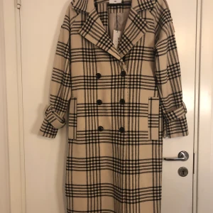 NA-KD kappa - Mysig, oanvänd kappa från NA-KD. Orginalpris 1000 kr. Beige med svarta ränder och svarta knappar. Silver detaljer på skärp och armar. strl 34